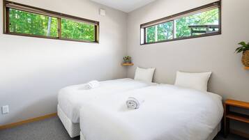 A hotel room with two beds, a small shelf with a plant, and windows with a view of trees.