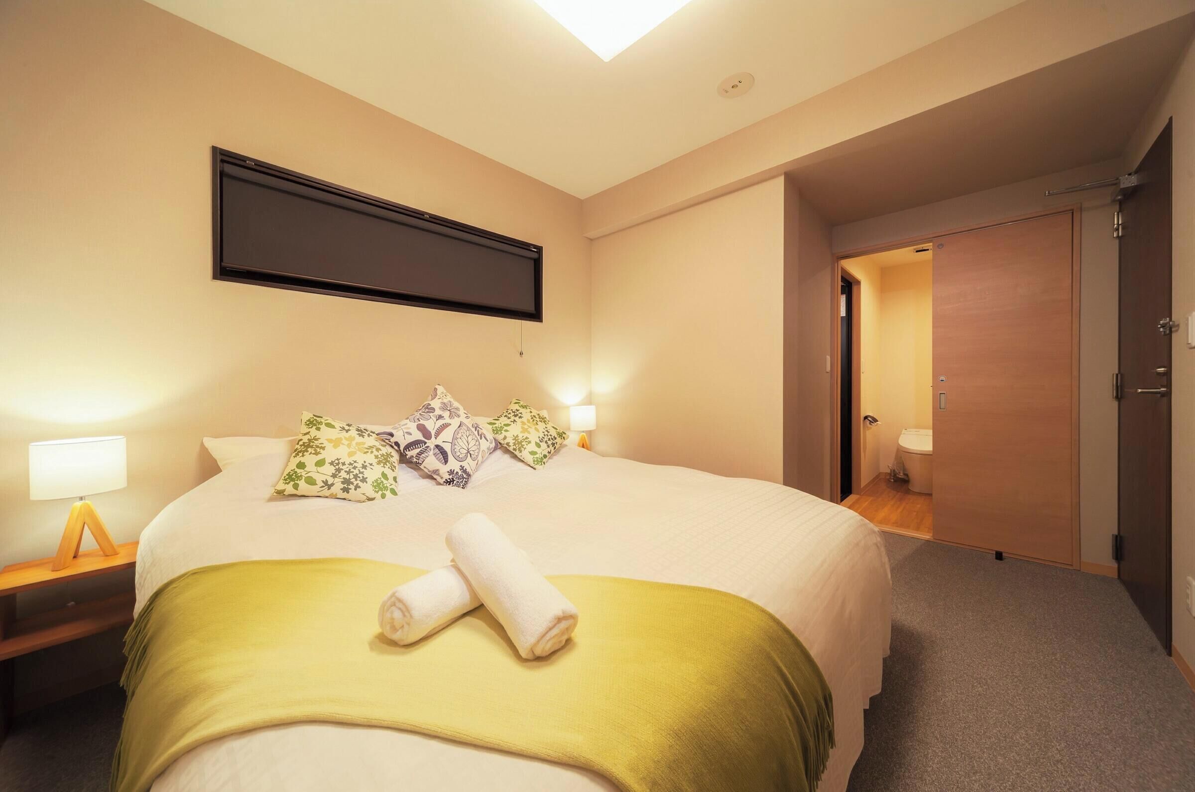 Quarto de hotel com cama, televisão de tela plana na parede, mesa de cabeceira com abajur e porta de acesso ao banheiro.