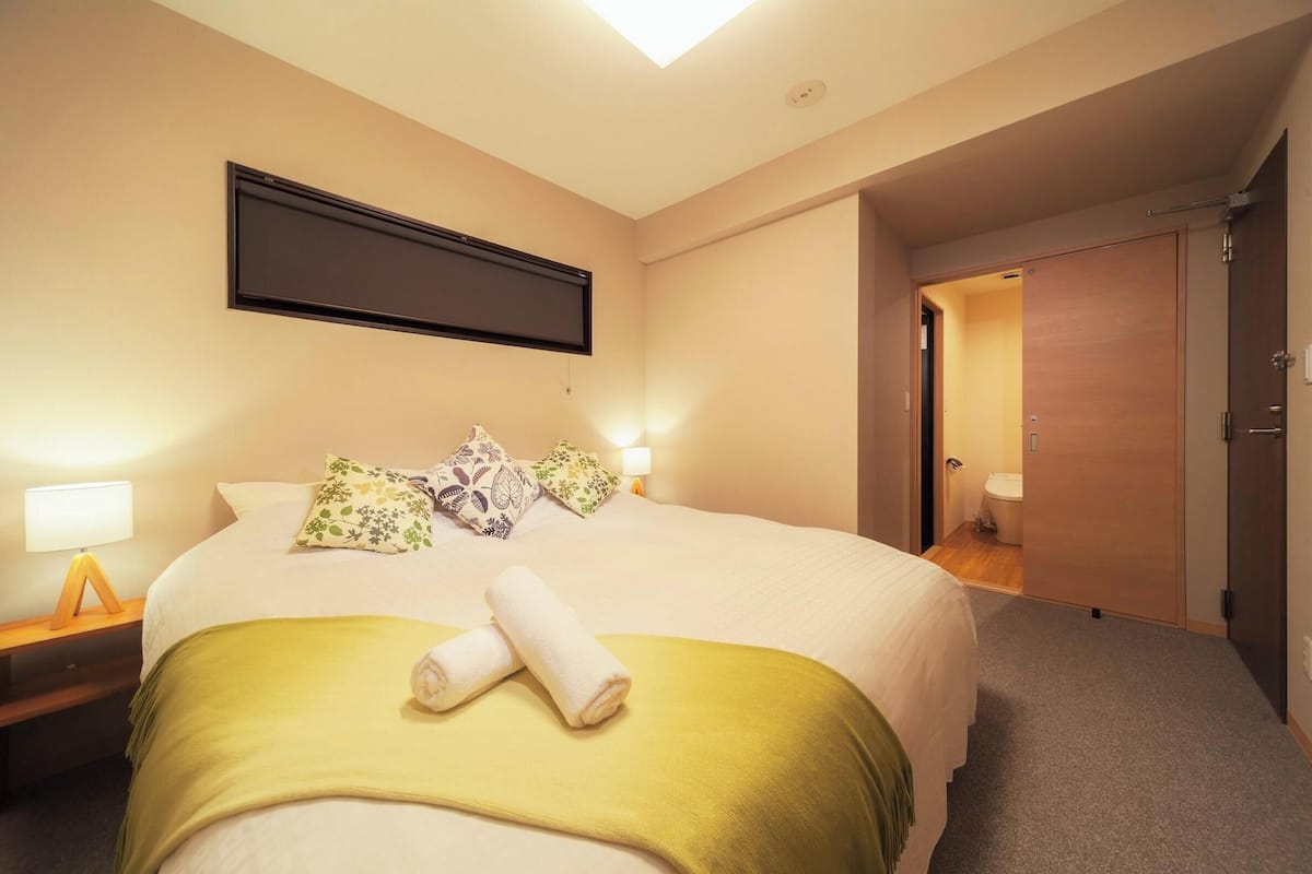 A hotel room with a bed, a flat-screen TV mounted on the wall, a bedside table with a lamp, and a door leading to a bathroom.