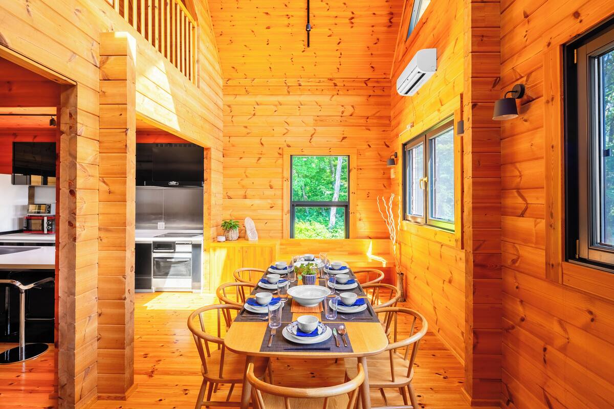 A wooden cabin interior with a dining table set for six, a kitchen area, and a window with a view of greenery.