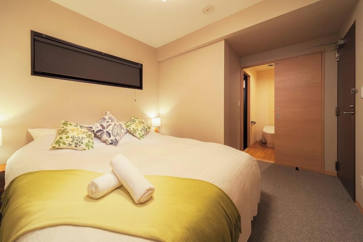 A hotel room with a bed, a TV mounted on the wall, a bedside lamp, and a door leading to a bathroom.