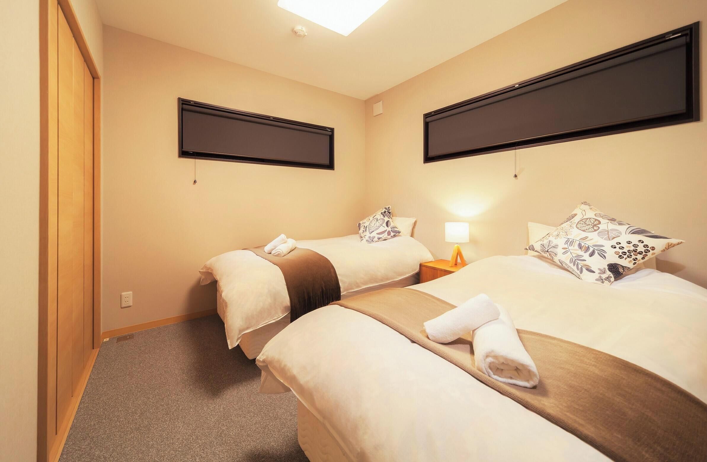 Quarto de hotel com duas camas, uma televisão de tela plana na parede, uma mesinha de cabeceira com abajur, e uma toalha sobre cada cama.
