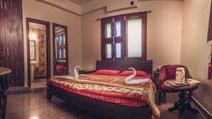 A spacious bedroom with a large bed, a sitting area with a chair and table, and a bathroom visible through an open door. - Kalyan Heritage (Jaipur)