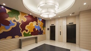Un vestíbulo de hotel con una pared floral de colores, una lámpara moderna y dos ascensores.