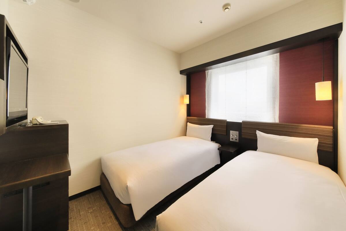A hotel room with two single beds, a desk with a telephone, and a window with a view.
