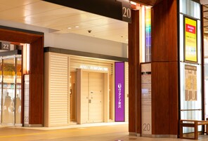 Entrada de hotel con una persiana cerrada, un letrero vertical morado y un banco de madera en el exterior.