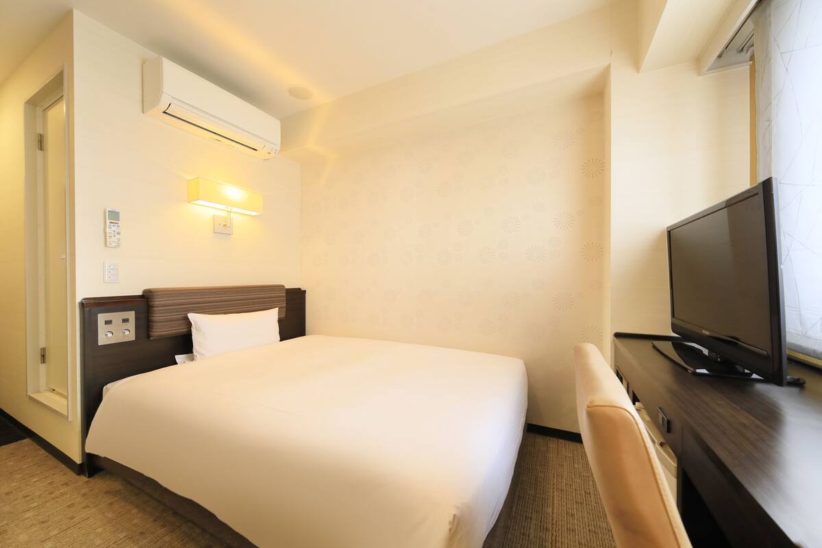 A hotel room with a bed, a desk with a television, an air conditioner, and a wall-mounted light fixture.