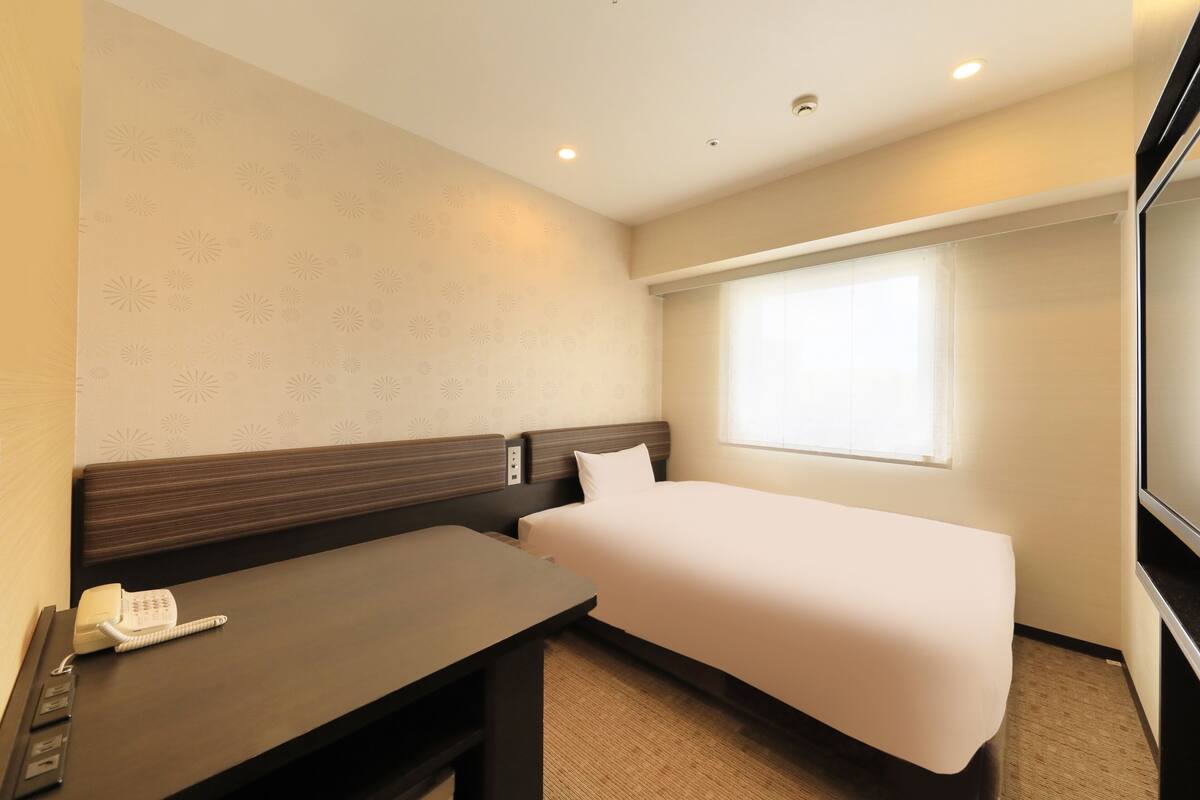 A hotel room with a single bed, a desk with a telephone, and a window with curtains.
