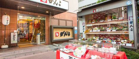 Um carrinho de comida de rua com diversos pratos expostos em uma vitrine, um aviso indicando uma promoção especial e uma fachada tradicional japonesa ao fundo.
