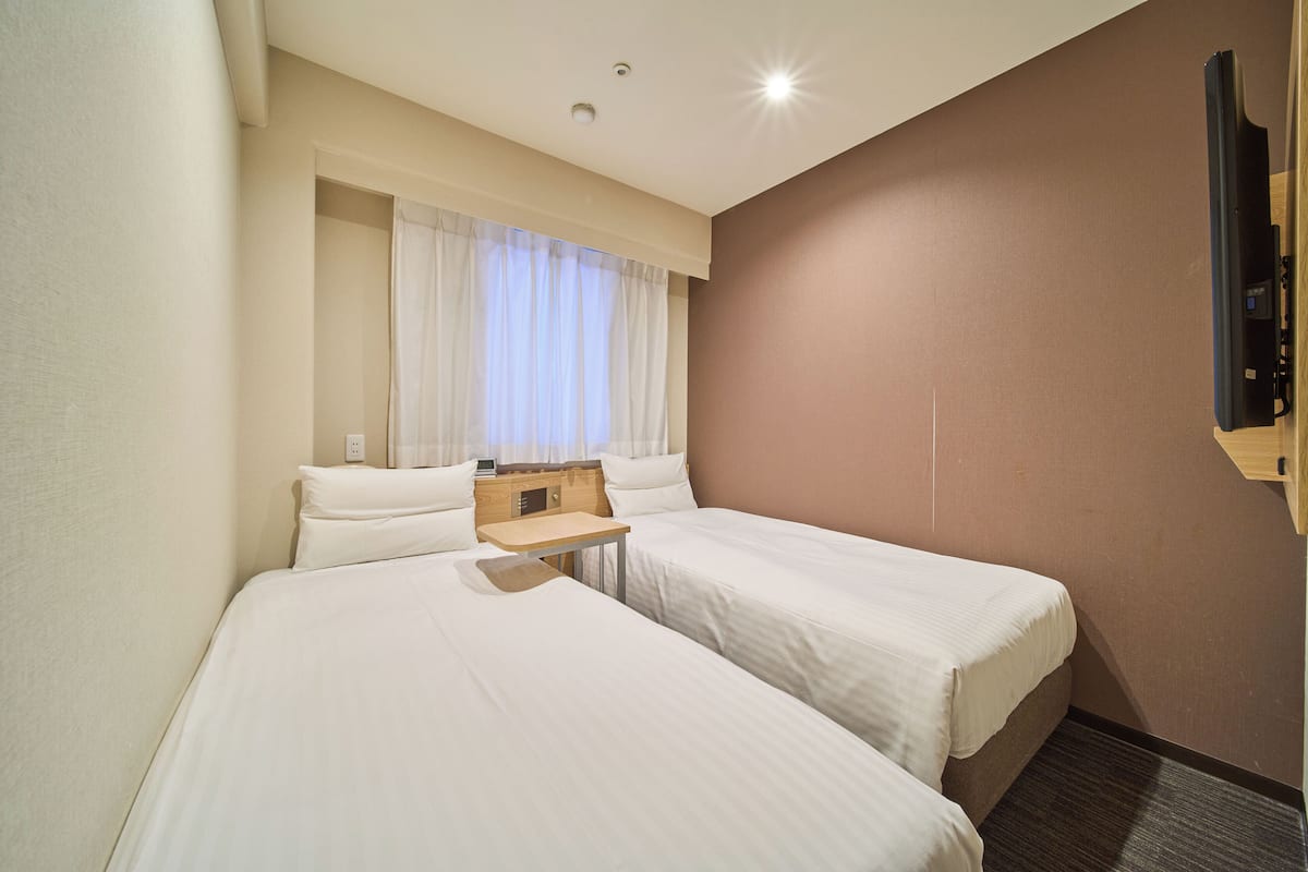 Una camera d'albergo con due letti, un tavolino, una TV a parete e una finestra con tende.