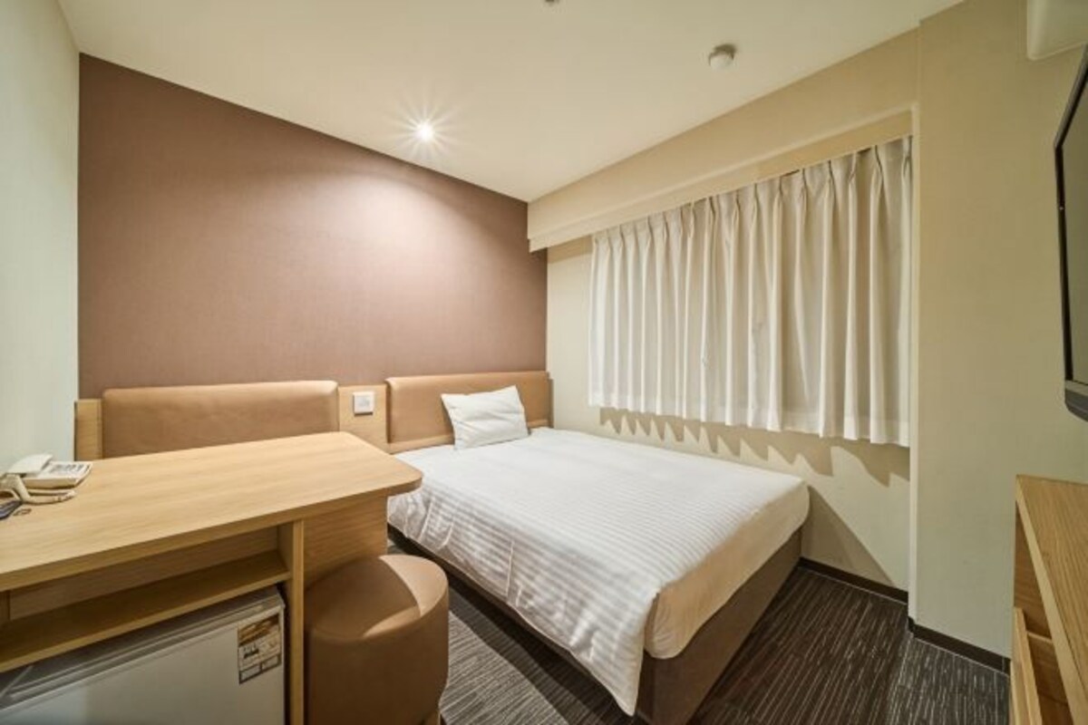 Une chambre d’hôtel avec un lit, un bureau, une chaise, une petite table et une fenêtre avec des rideaux.