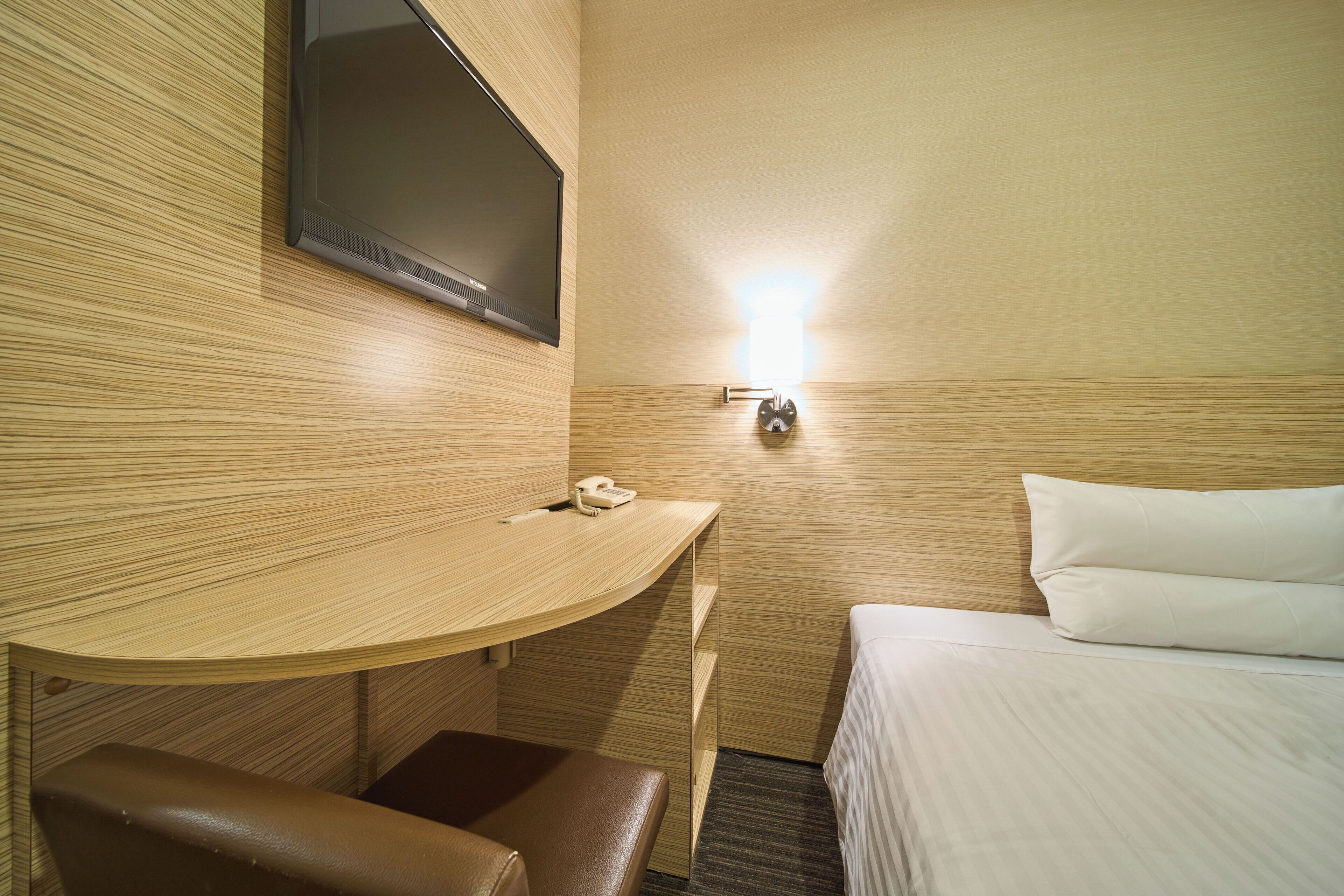 A hotel room with a wooden wall, a flat-screen TV mounted above a desk, a bedside table with a phone, and a bed with white bedding.