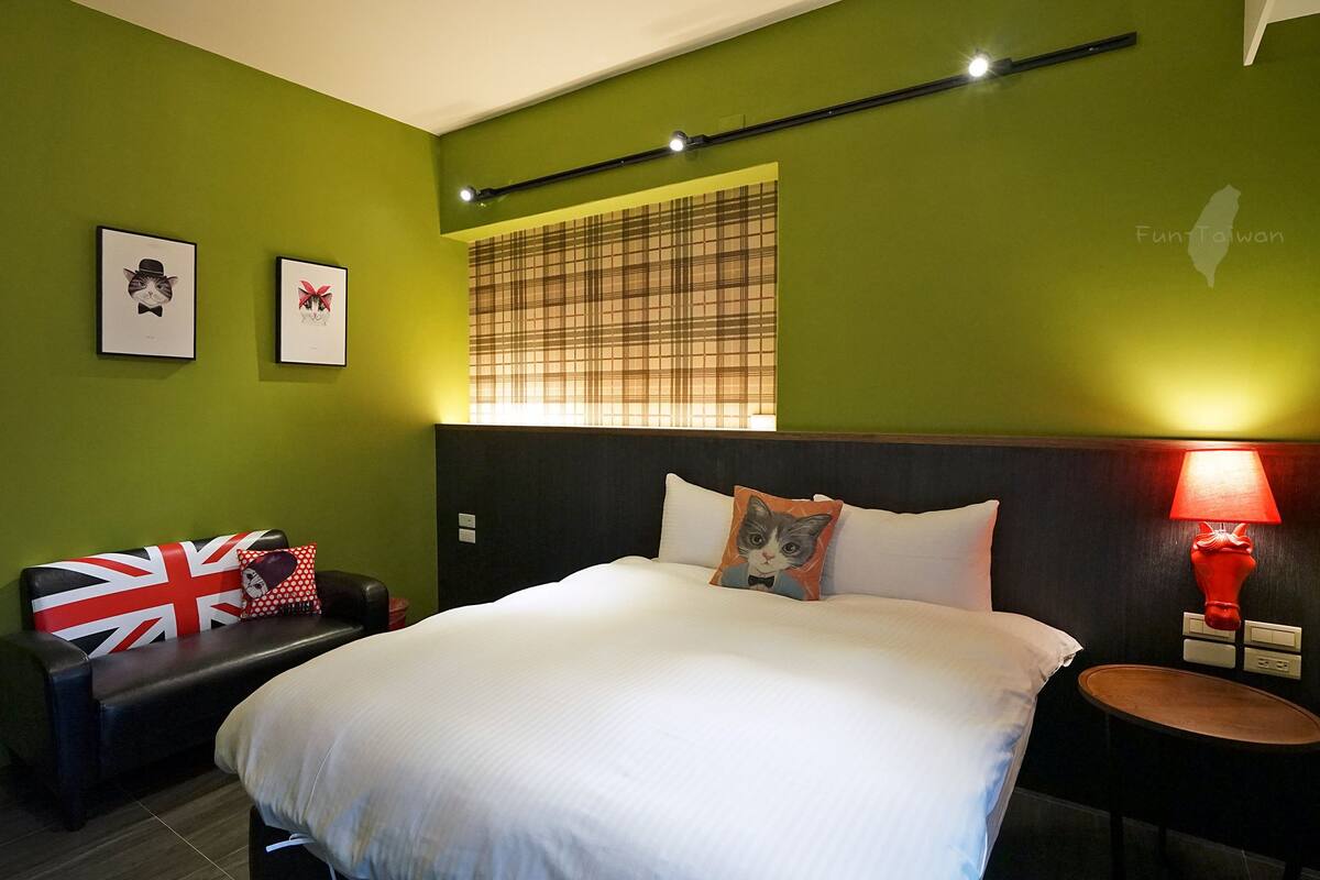 Una habitación de hotel con una cama, una mesita de noche con una lámpara, una silla con un cojín con la bandera Union Jack y dos cuadros en la pared.