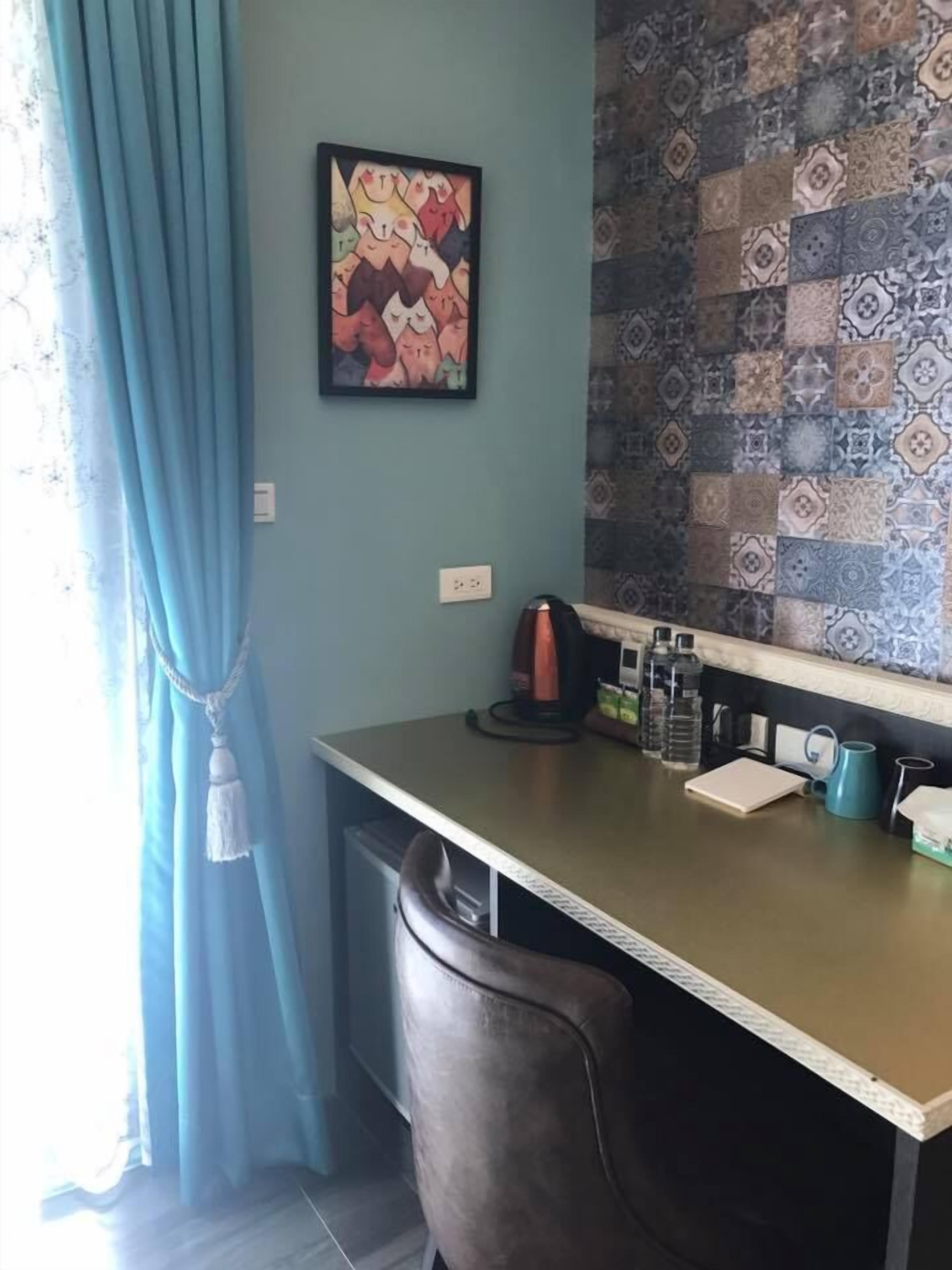 A room with a desk, a chair, a wall with patterned tiles, a painting, and a window with curtains.