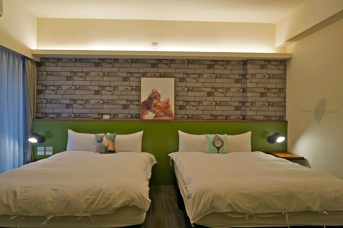 Habitación de hotel con dos camas, pared de ladrillo, un cuadro enmarcado de un gato y una pared de acento verde.