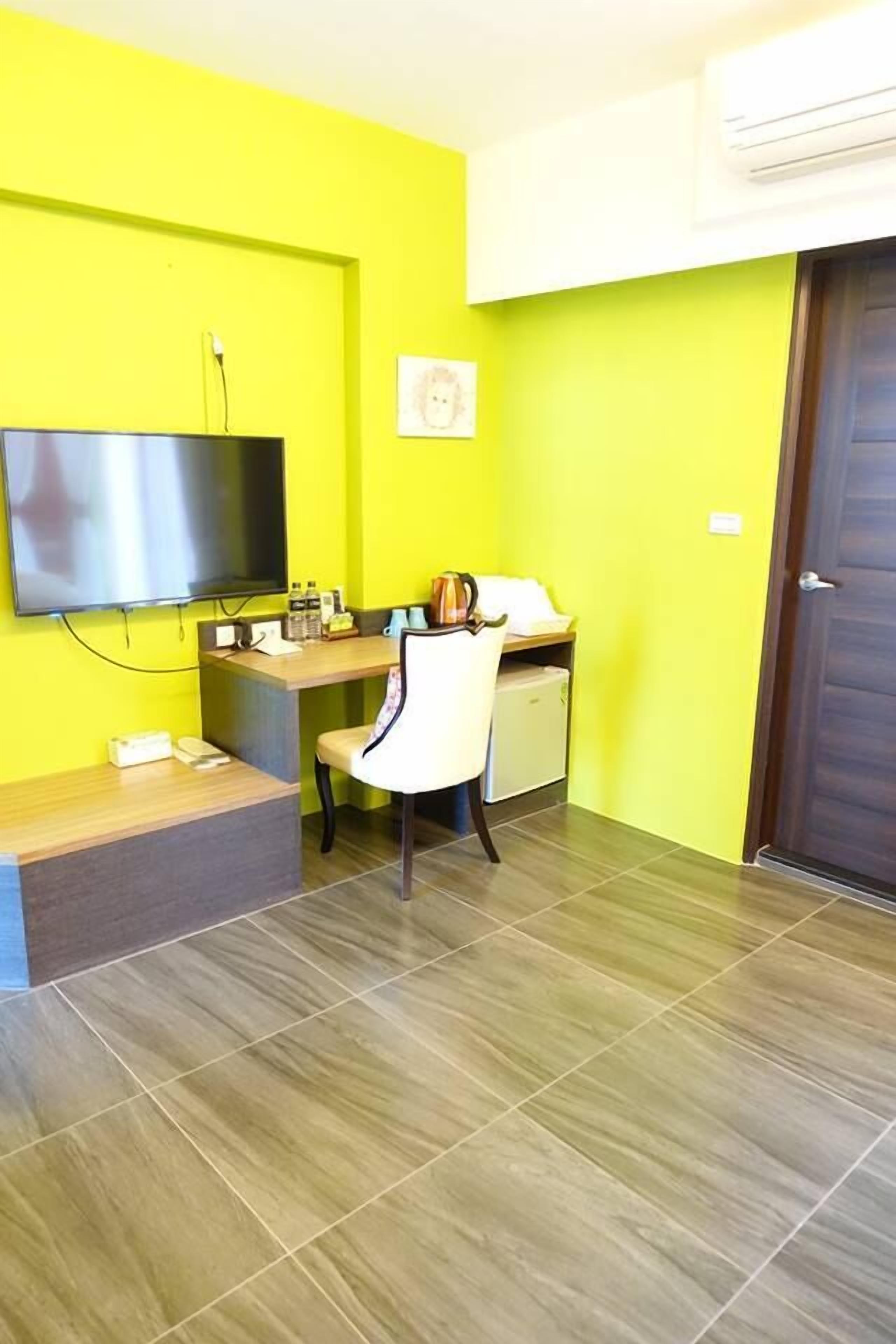 A room with a bright yellow wall, a flat-screen TV mounted on the wall, a desk with a chair, and a door.