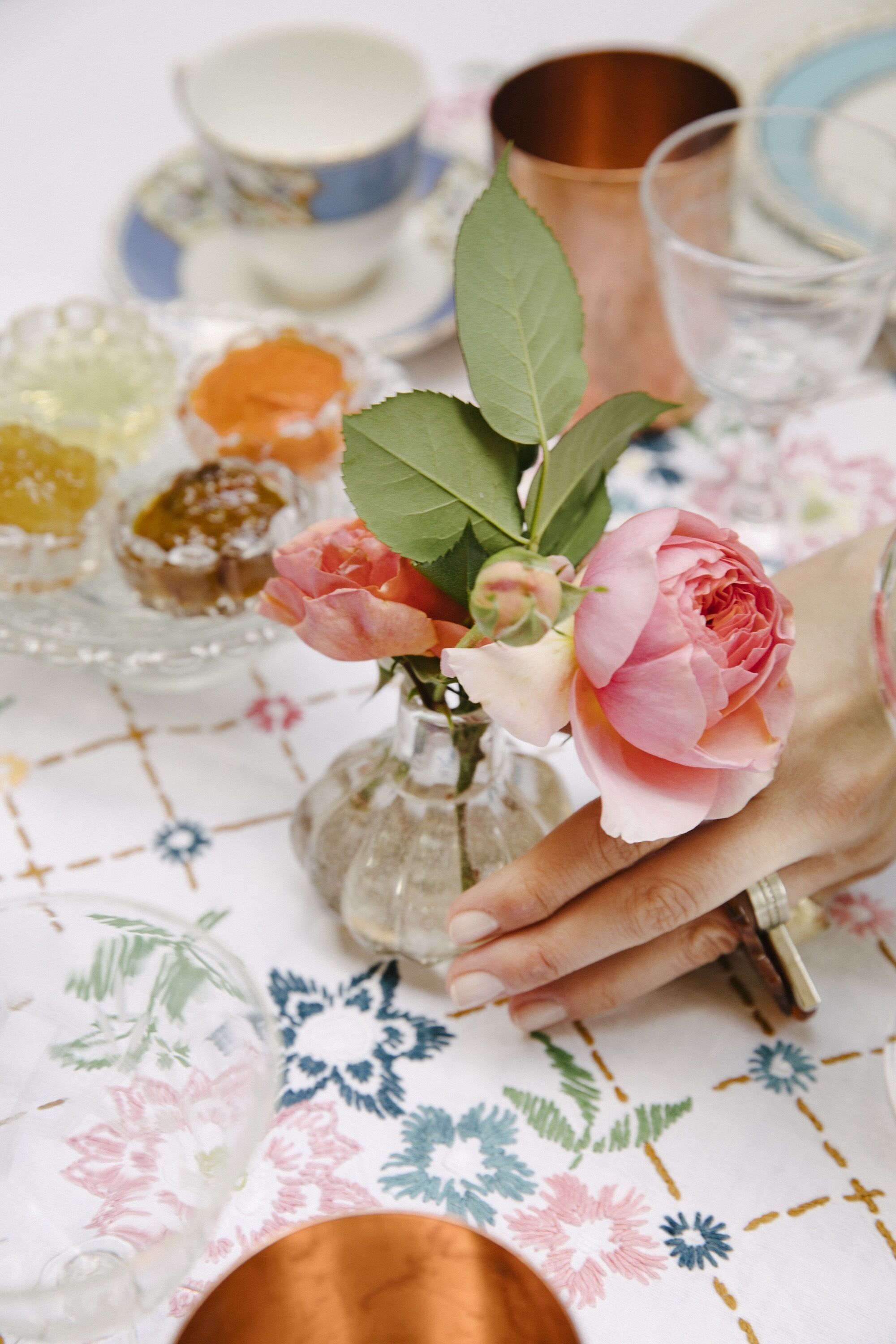 Uma mesa com toalha florida, um vaso com rosas cor-de-rosa e diversos copos e pratos.