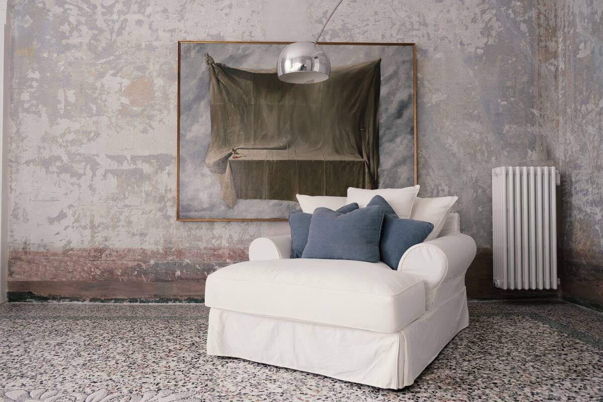 Un sofá blanco con cojines azules en una habitación con pared de concreto texturizado y un radiador.