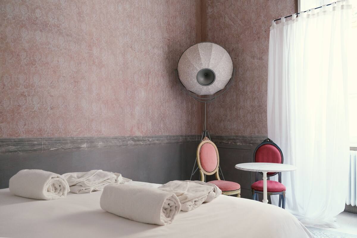 Una habitación de hotel con una cama, dos sillas, una mesita y una lámpara fijada a la pared.