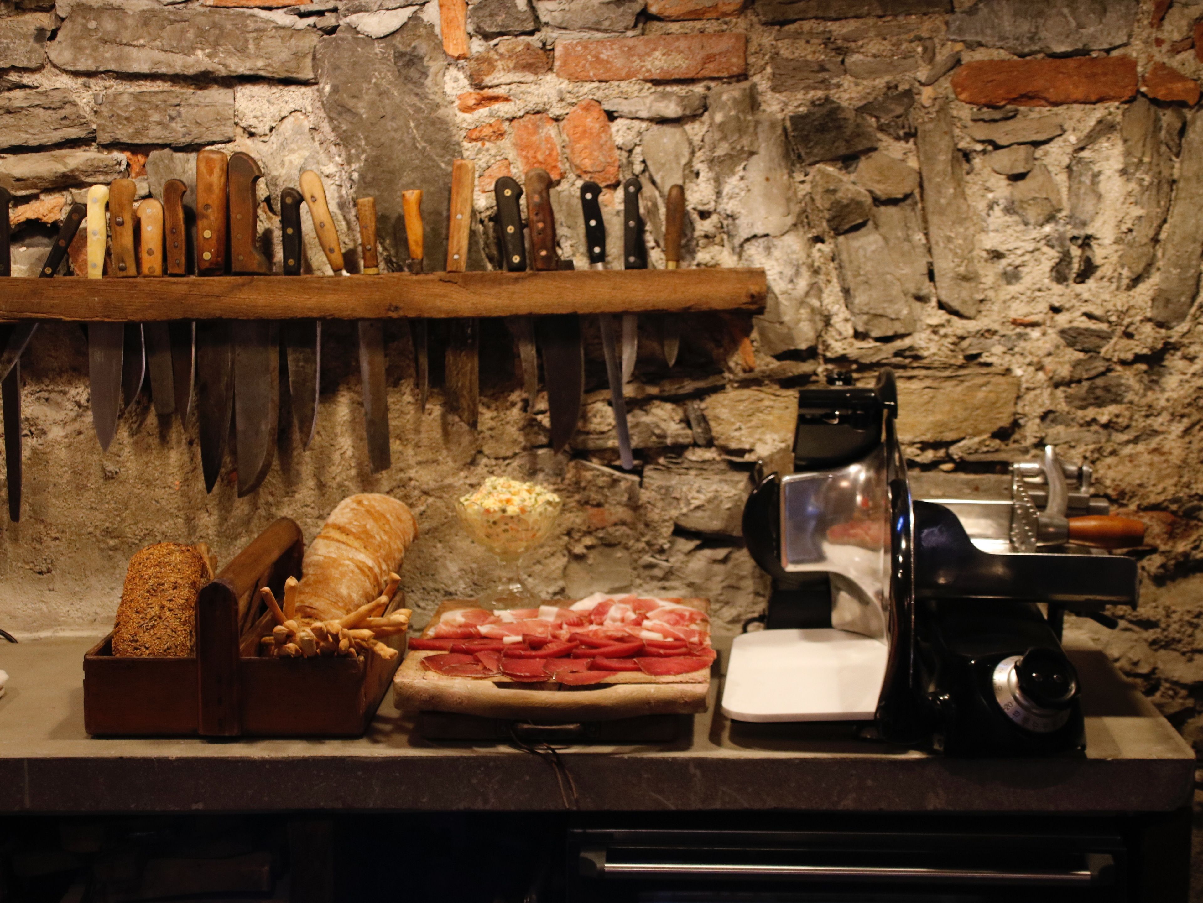 Una cocina con pared de piedra, repisa de madera con cuchillos, un cortador de carne y una cesta de pan.