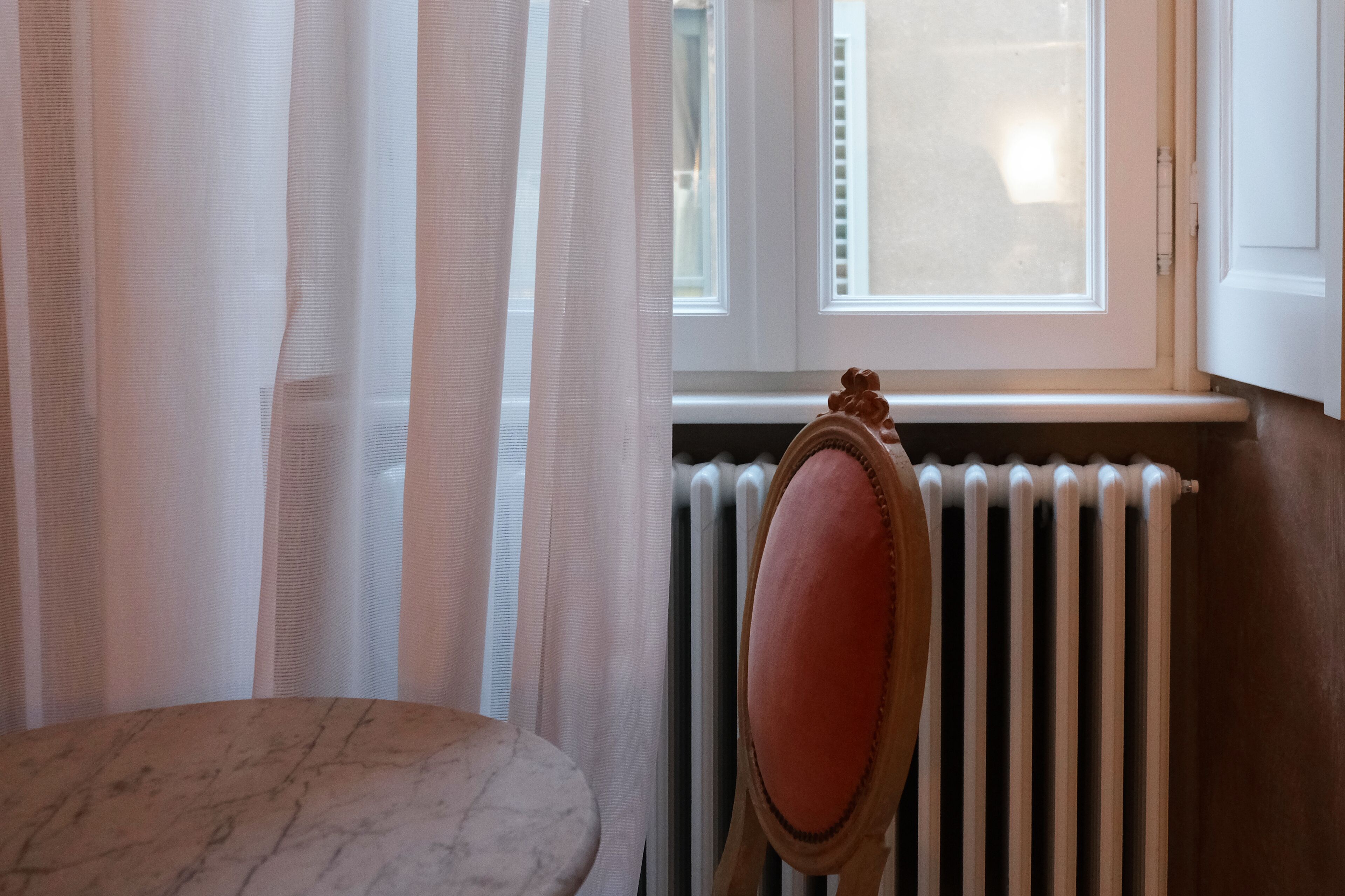 Una habitación con un radiador, una silla con cojín y cortinas traslúcidas.
