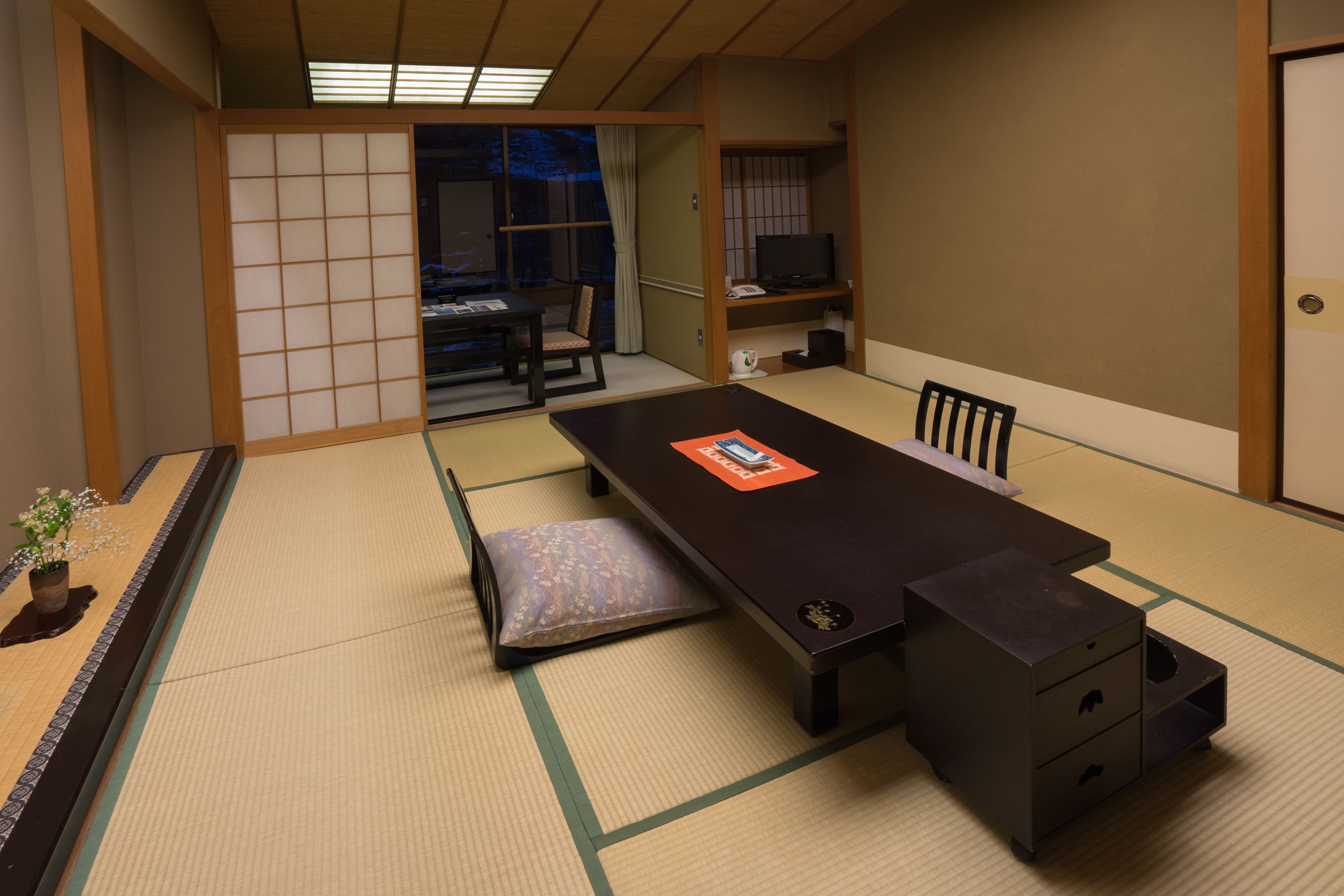 A traditional Japanese room with tatami flooring, a low table, a chair, a television, and a sliding door leading to another room.