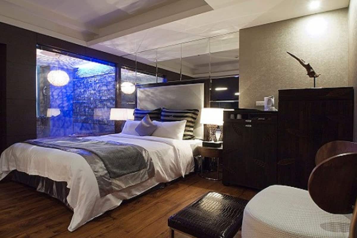 A modern hotel room with a large bed, a desk, a wardrobe, and a view of a cityscape through a large window.