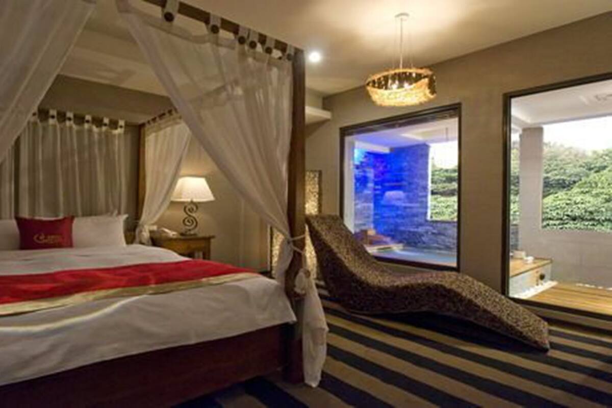 A bedroom with a bed, a nightstand, a lamp, a chaise lounge, and a view of greenery outside.