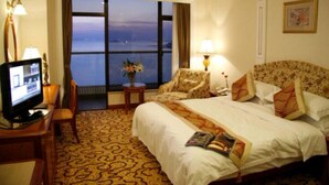 A hotel room with a large bed, a television, a small table with a lamp, and a view of the sea through a large window. - Huiquan Dynasty Hotel (Qingdao)