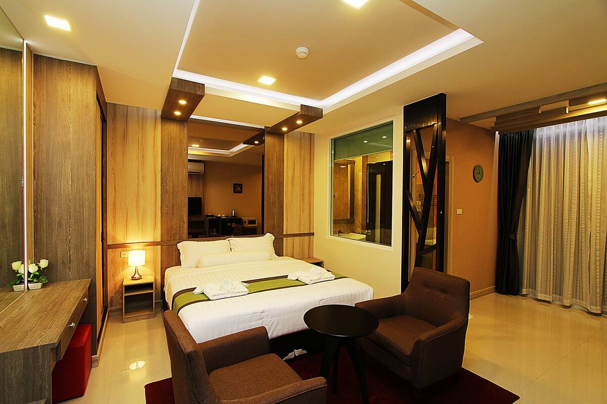 Une chambre d’hôtel avec un grand lit, un coin salon avec deux fauteuils, une petite table, un miroir et une fenêtre avec des rideaux.