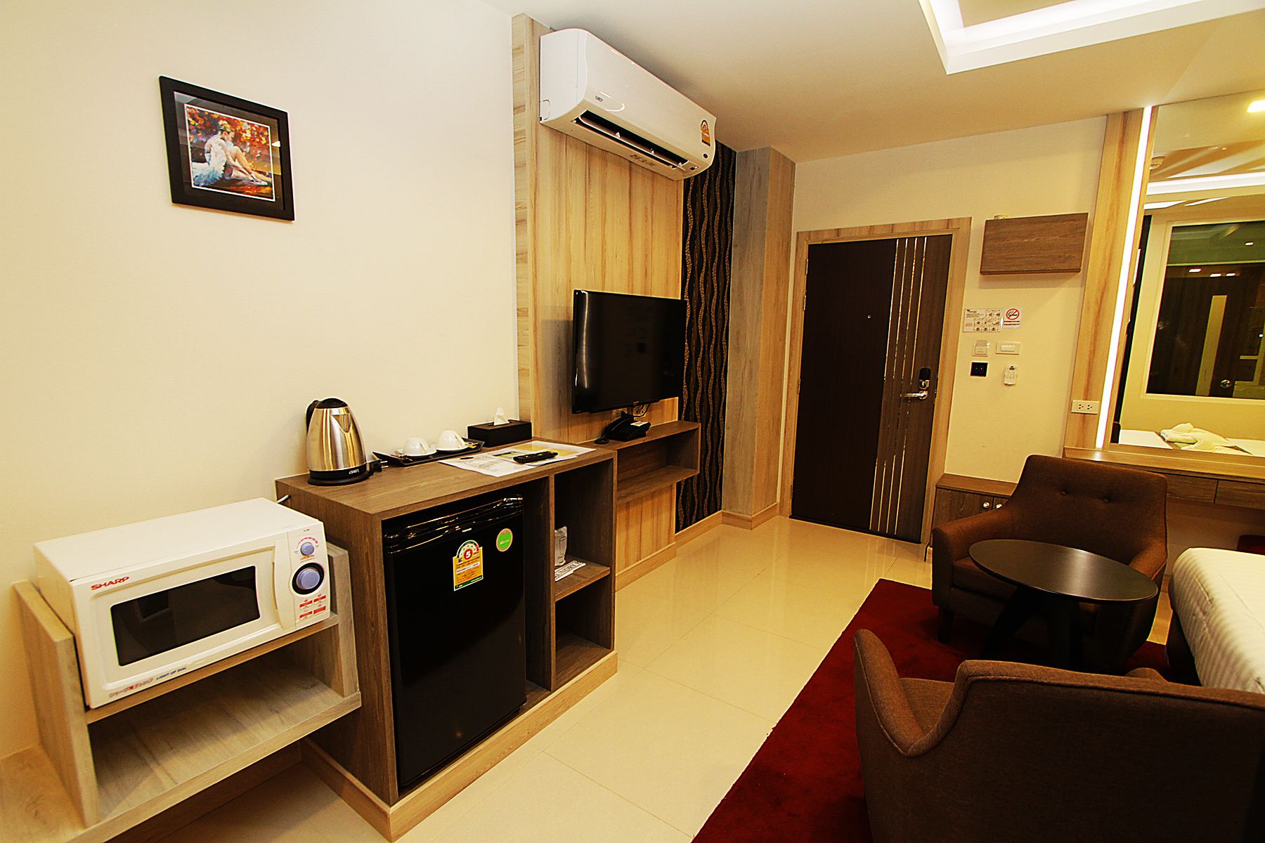 A compact hotel room with a kitchenette, a flat-screen TV, a microwave, a refrigerator, a wooden cabinet, a sofa, and a red carpet.