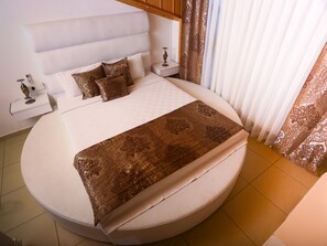 Una cama redonda con cabecera blanca, cubrecama marrón estampado y almohadas a juego. Hay dos mesitas de noche, una con lámpara y un objeto decorativo pequeño.
