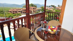 Un balcón con sillones de mimbre, una mesa con frutero y copas de vino, y vistas a casas y montañas.