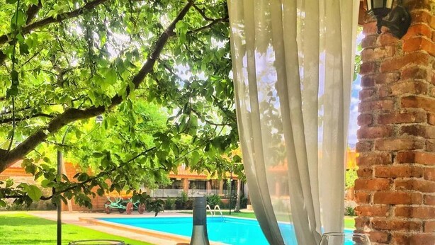 A table with a fruit platter, two glasses of red wine, and a bottle of sauce, set against a backdrop of a swimming pool and lush greenery. - Mogla Tashan Boutique Otel (Mugla)