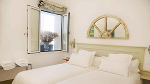 A double bed with white linens, a bedside table with a lamp, and a window with a view of a building and trees.