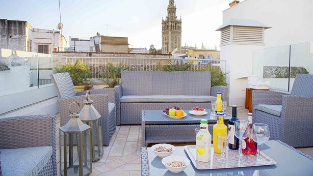 A rooftop terrace with wicker furniture, a tray with food and drinks, and a view of a historic tower in the background.