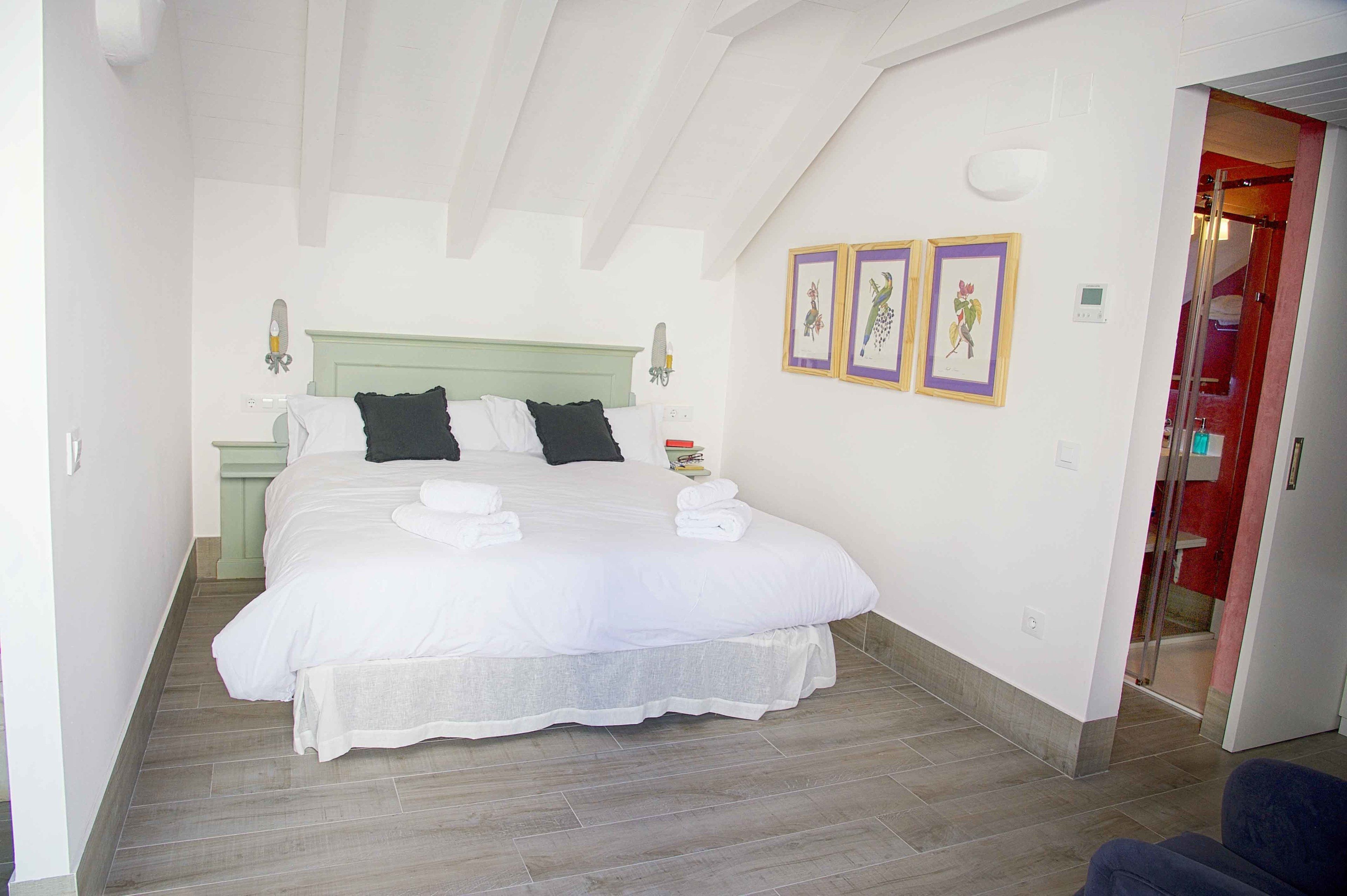 Una cama bien hecha con sábanas y almohadas blancas, una mesita de noche con una lámpara y una pared con tres cuadros enmarcados.