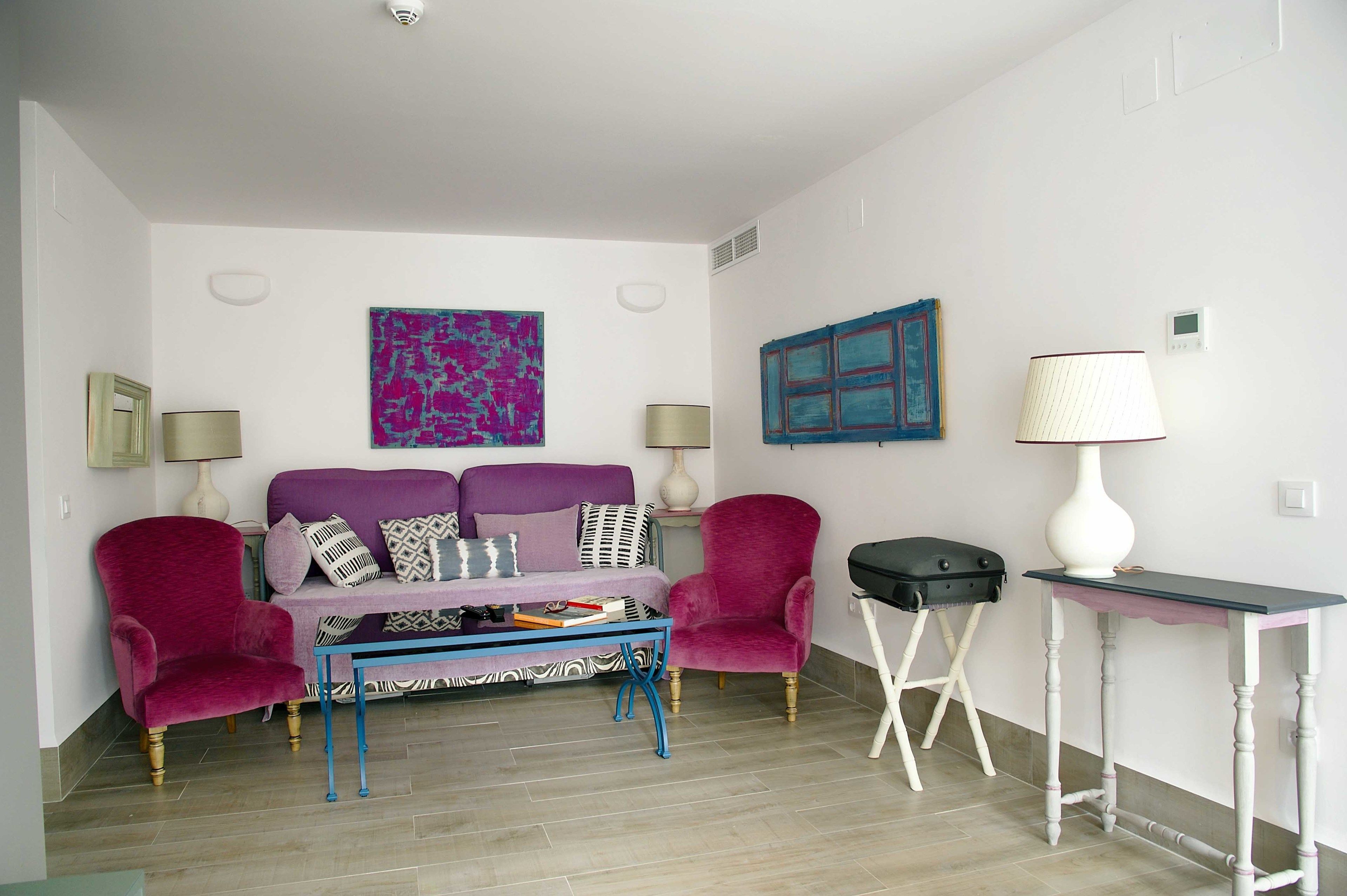 Una habitación con un sofá morado, dos sillones, una mesita de centro, una lámpara y un cuadro en la pared.