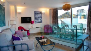 A modern living room with a sofa, a glass coffee table, a red armchair, a TV, and a balcony with a table and chairs.