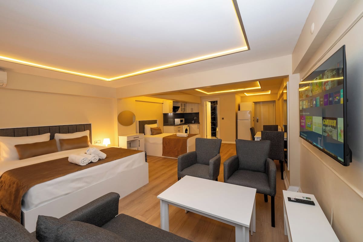 A hotel room with a bed, two armchairs, a small table, a TV, and a kitchenette in the background.
