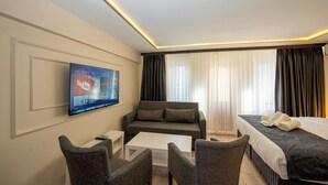 Habitación de hotel con televisor de pantalla plana en la pared, sofá, cama con cabecera negra, mesita de noche blanca y una pequeña mesa blanca.