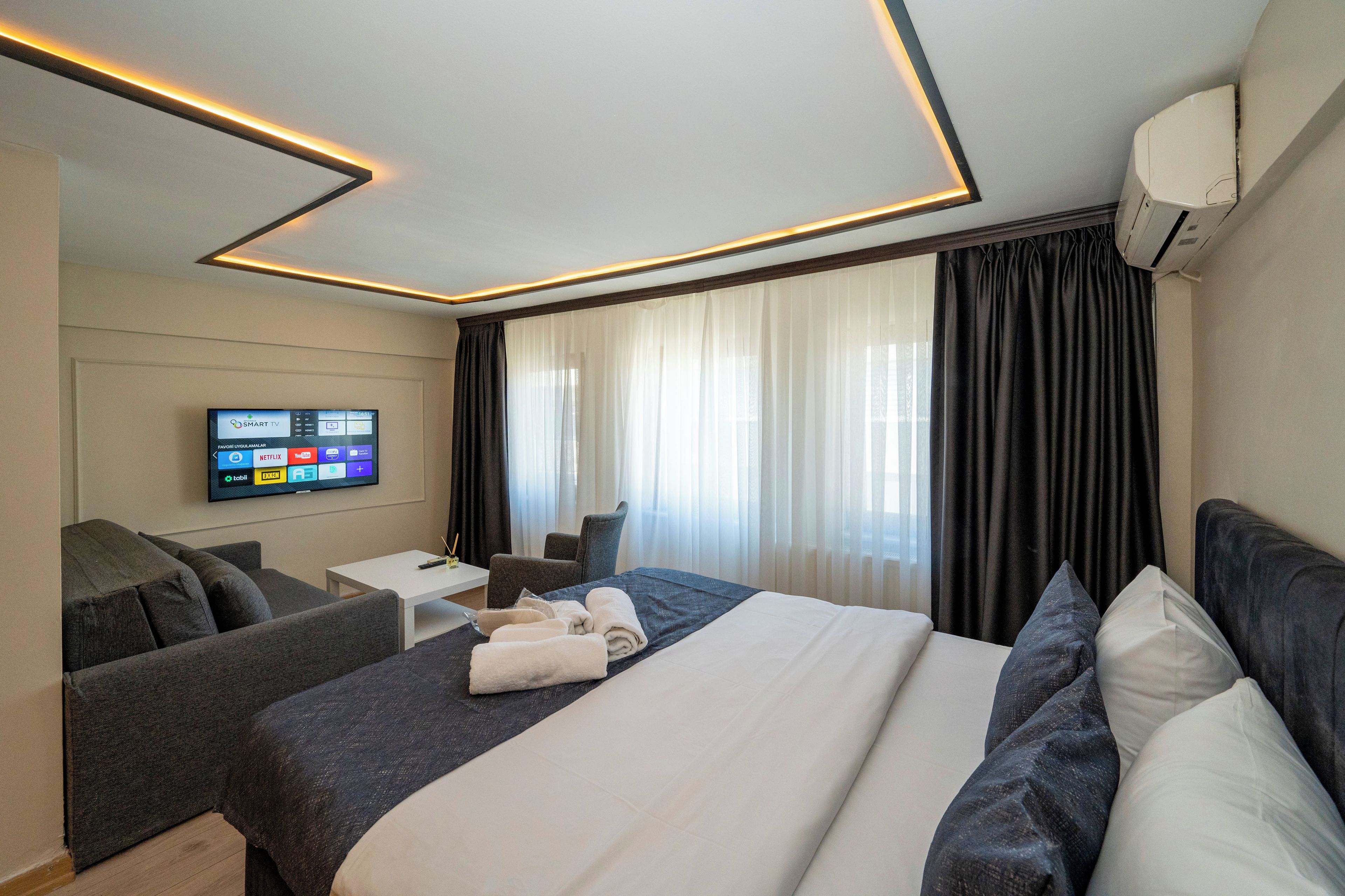 A modern hotel room with a large bed, a flat-screen TV mounted on the wall, a seating area with a small table, and a large window with curtains.