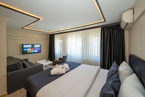 A modern hotel room with a large bed, a flat-screen TV mounted on the wall, a seating area with a small table, and a large window with curtains. - Aiza Suites (Istanbul)