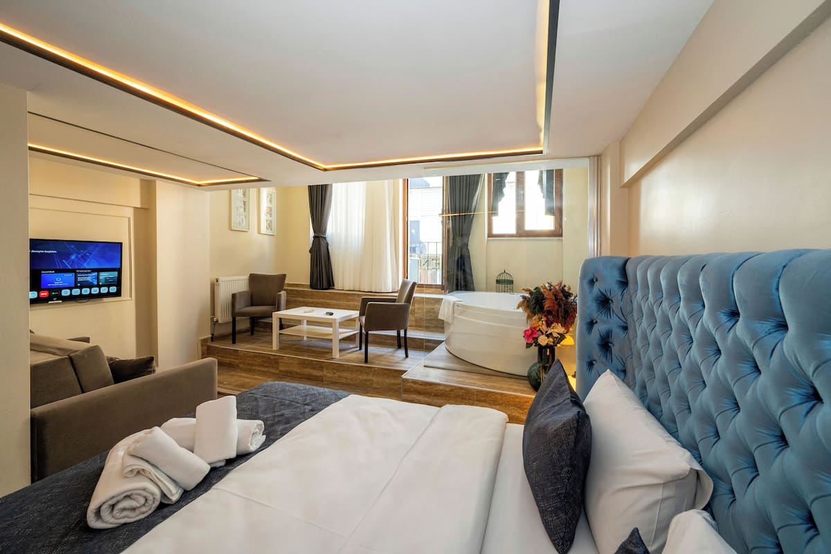 A modern hotel room with a large bed, a flat-screen TV, a seating area with a sofa and chairs, and a view of the city.