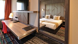 A hotel room with a wooden desk, a flat-screen TV mounted above it, a bed with a headboard featuring geometric patterns, and a colorful rug on the floor. - XO Hotels Couture (Amsterdam)