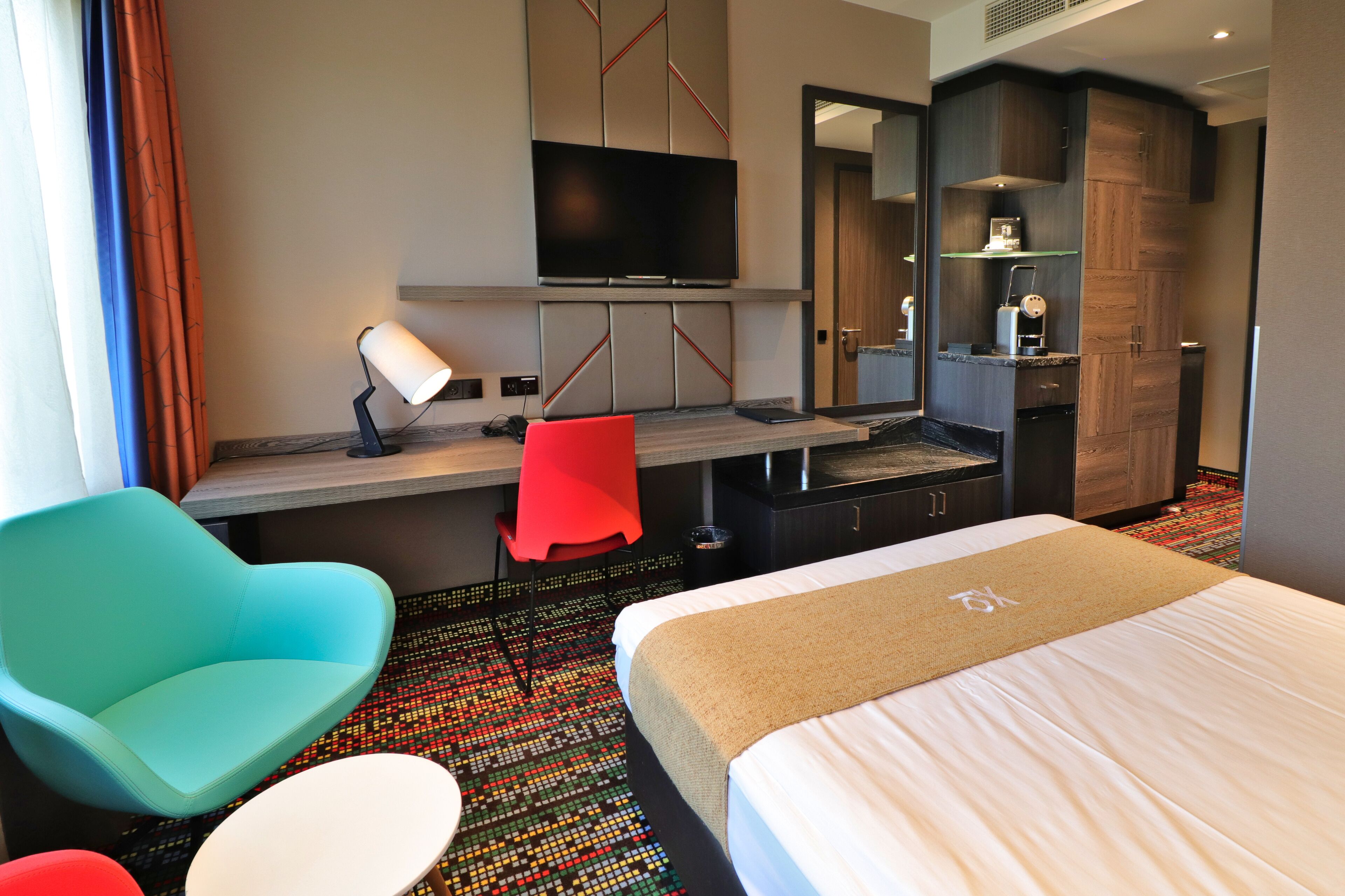 A hotel room with a bed, a desk with a TV, a red chair, and a small table.
