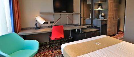 A hotel room with a bed, a desk with a TV, a red chair, and a small table.