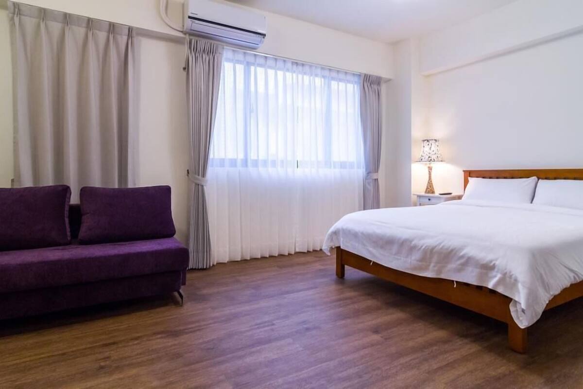 Une chambre d’hôtel avec un plancher en bois, un lit avec une literie blanche, un canapé violet et une fenêtre avec des rideaux.