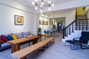 Een ruime woonkamer met een houten bank, een bank met kleurrijke kussens, een eettafel en stoelen. Er is een trap die naar een bovenverdieping leidt.