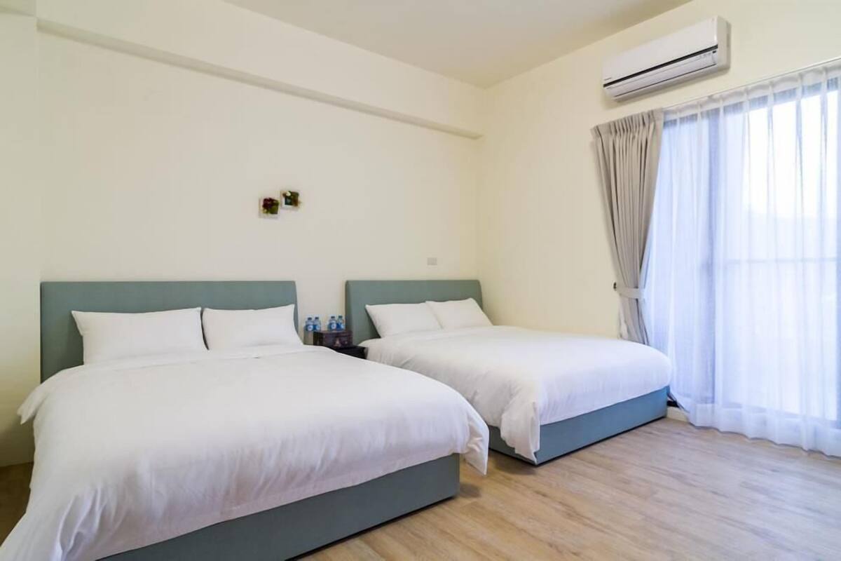 Une chambre d’hôtel avec deux lits simples, une petite table avec des bouteilles d’eau et une fenêtre avec des rideaux légers.